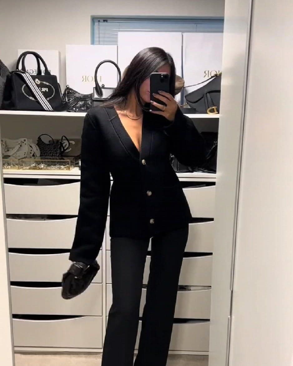 A mirror photo of an all-black outfit: a fitted knitted cardigan with gold buttons over black flared trousers and pointed heels