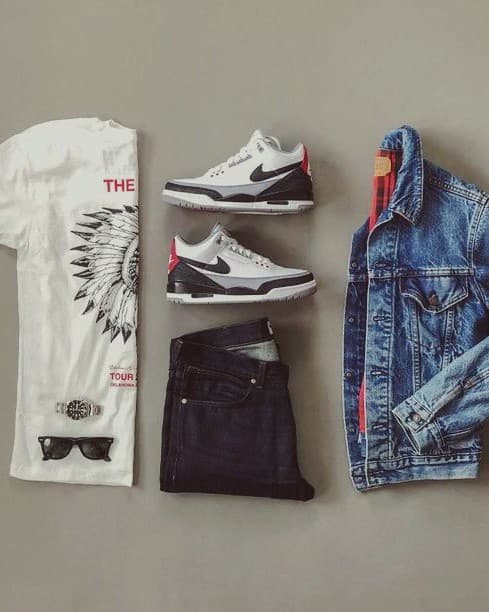 A flatlay of an outfit laid out on a grey floor: a white printed t-shirt, sunglasses, a watch, folded dark jeans, white and black sneakers and a denim jacket