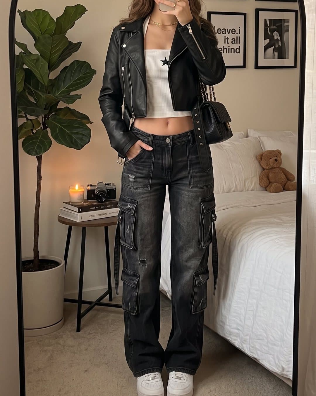 A mirror photo of an outfit made up of a cropped black leather biker jacket, a white star-print vest top, a small black shoulder bag and washed black cargo jeans