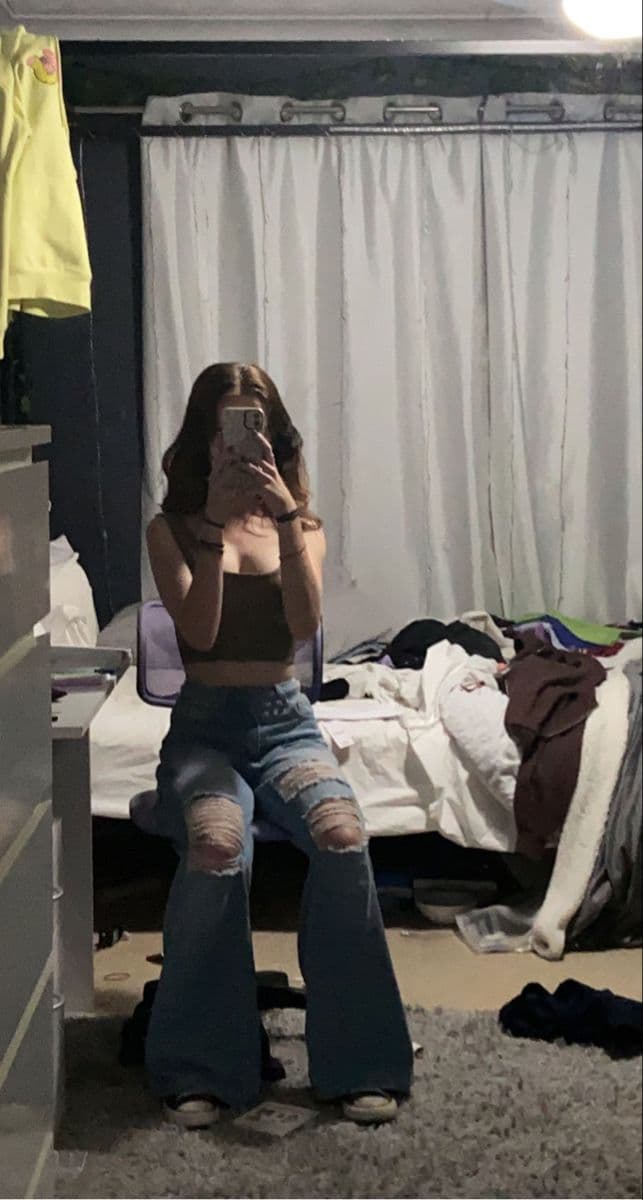 A bedroom mirror selfie with an unmade bed, piles of clothes and laundry on the floor behind, showing a black cropped vest top, ripped light blue wide-leg jeans and white trainers