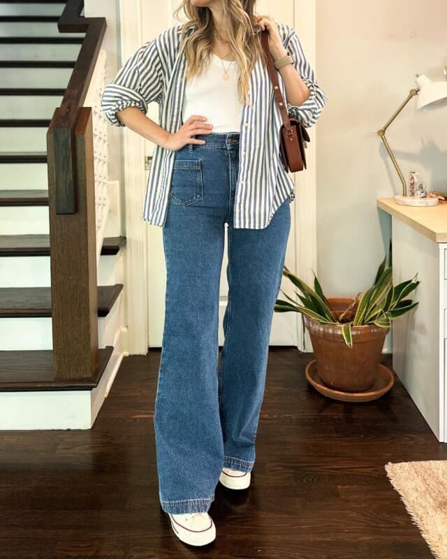A woman photographed head-on in an open blue striped overshirt, a white vest top, wide-leg blue jeans, a brown shoulder bag and white sneakers