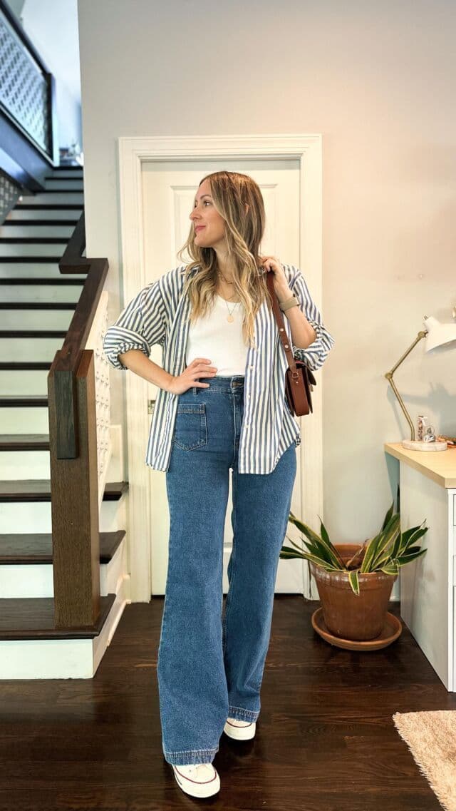A woman standing head-on in a hallway wearing an open blue striped shirt, a white vest top, wide-leg jeans, a brown shoulder bag and white sneakers, with the whole outfit visible from shoulders to shoes