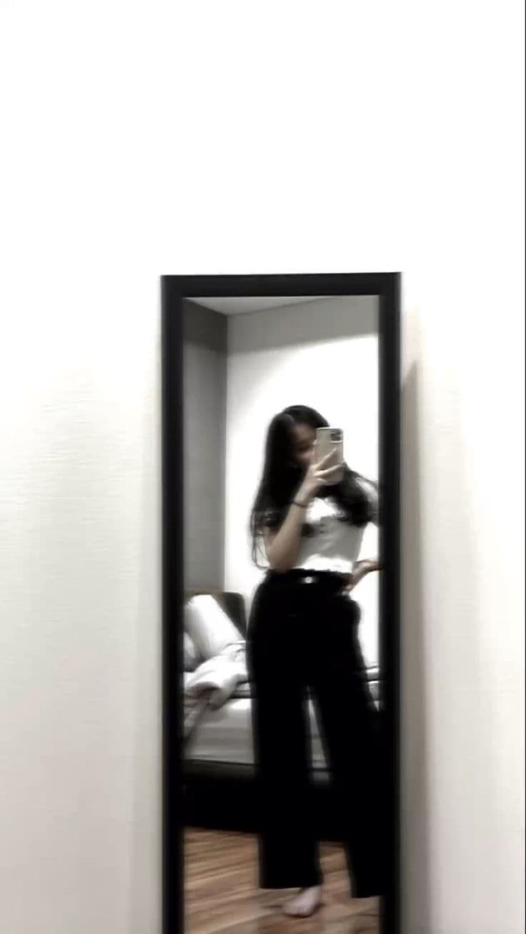 A dim mirror selfie taken from across a room, where the person and their outfit occupy a small part of a mostly empty white wall