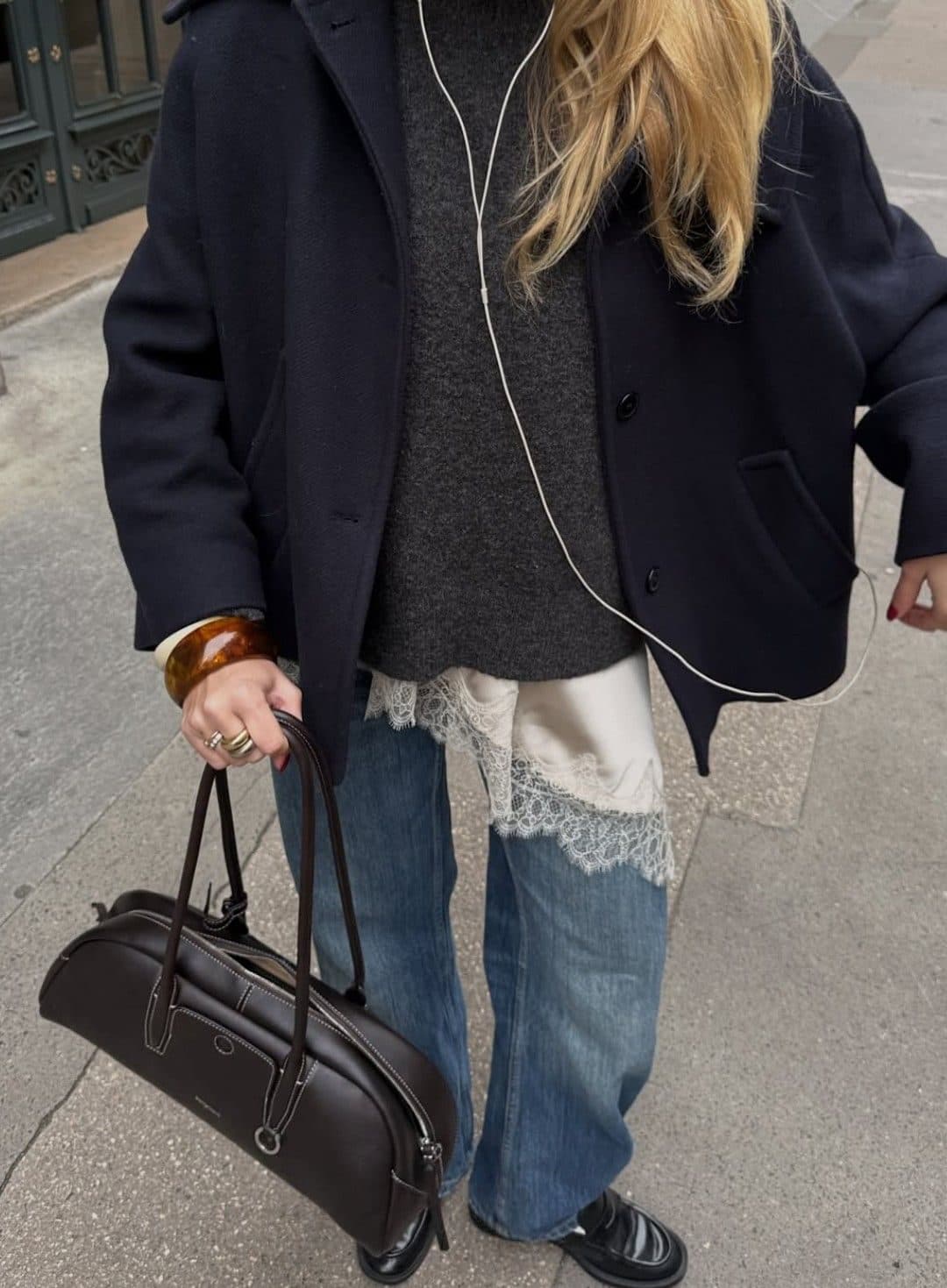 A cropped street shot of an outfit layering a navy wool coat over a charcoal jumper, a lace-hemmed slip and baggy jeans, with a brown leather holdall