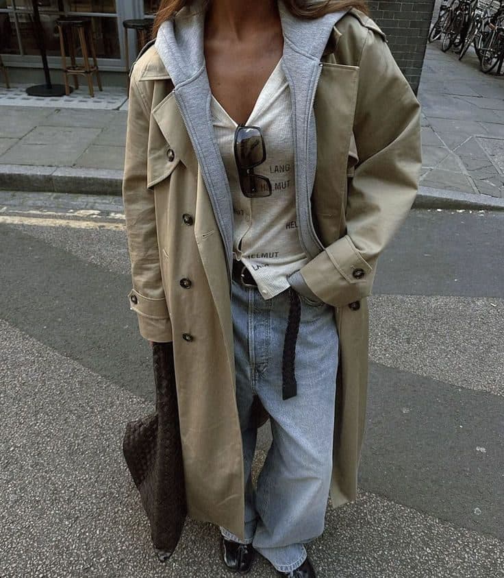 A cropped street shot of a stone trench coat worn open over a grey hoodie and a cream overshirt, with light blue baggy jeans