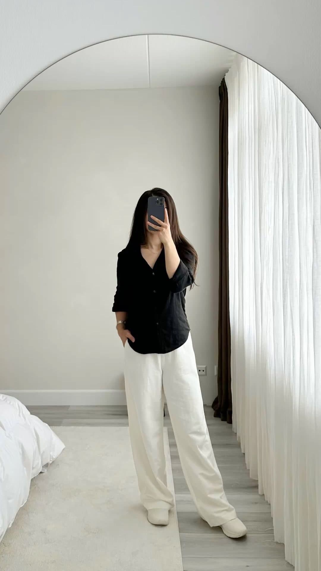 A full-length vertical frame of a woman standing still against a pale curved wall in a black shirt and cream wide-leg trousers