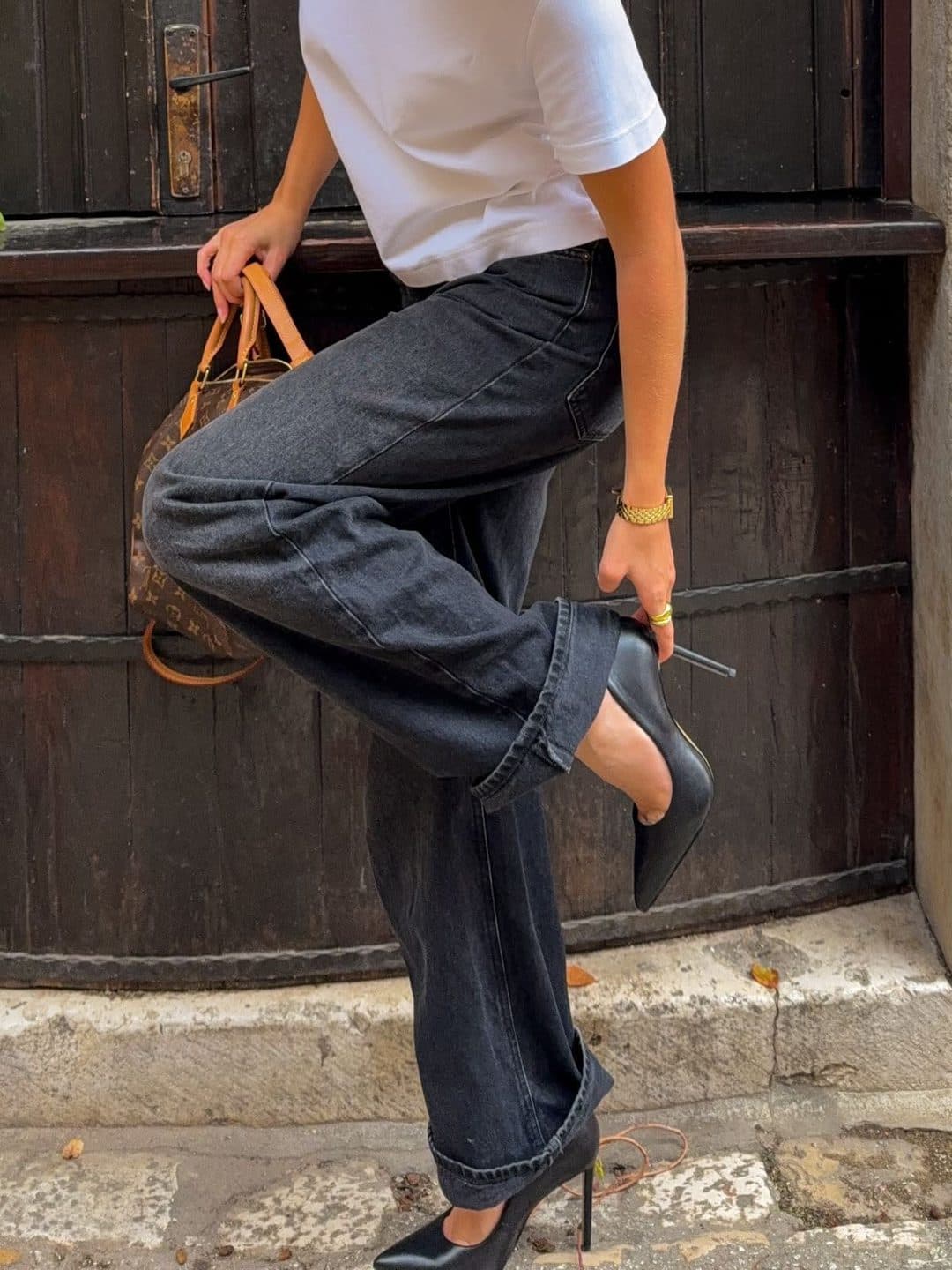 A cropped shot of dark wide-leg jeans and a black pointed slingback heel, one foot lifted against a wooden door