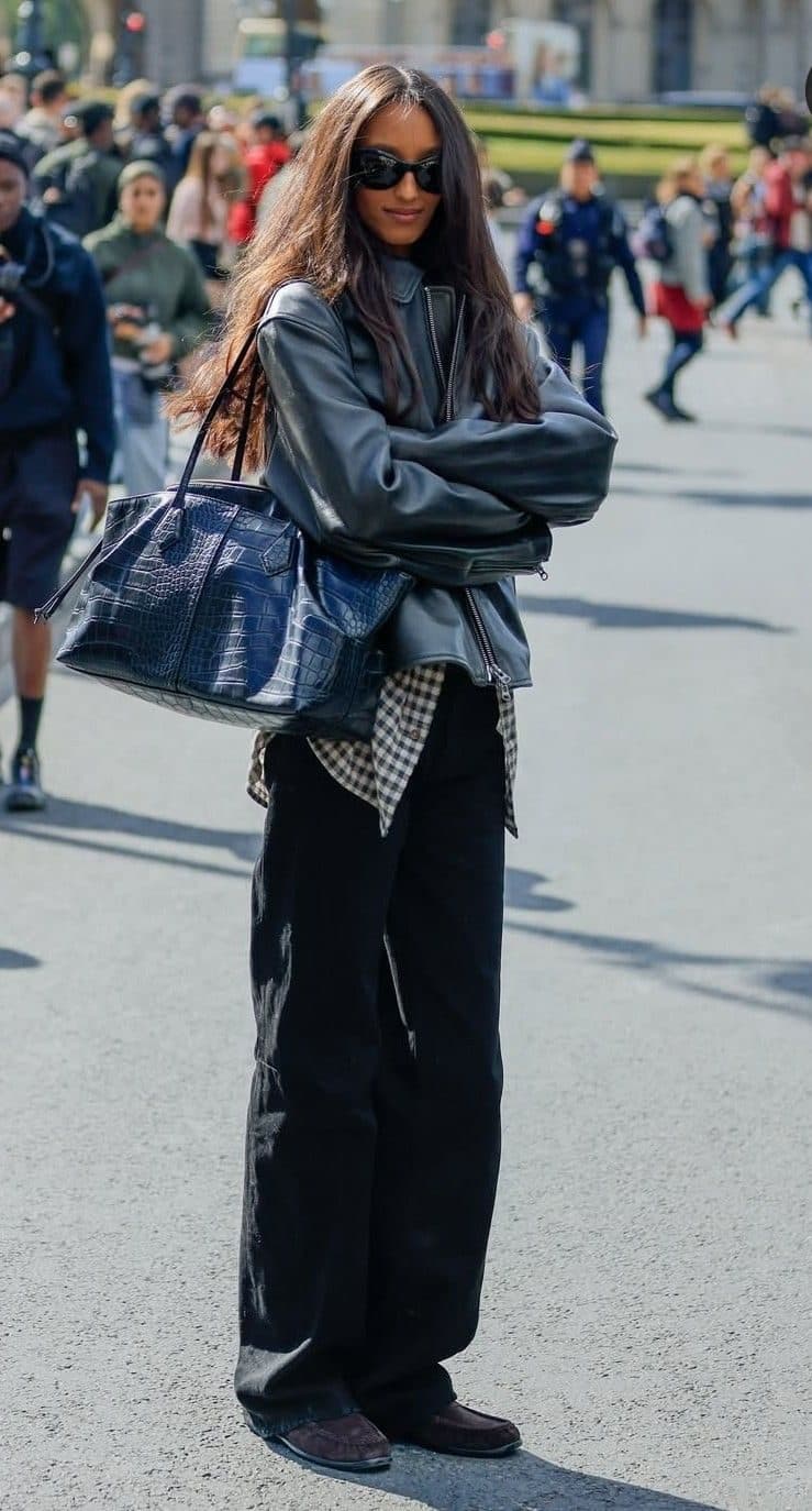 A woman in a grey leather jacket and black wide-leg trousers standing on a busy pavement with a crowd behind her