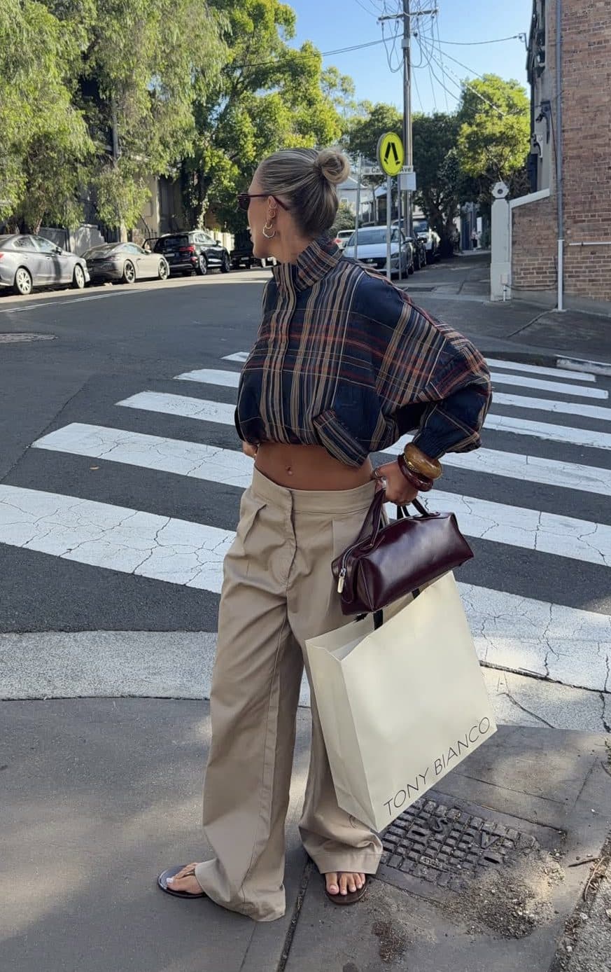 A woman crossing a street in a checked shirt, cream wide-leg trousers, flat sandals and a small burgundy bag