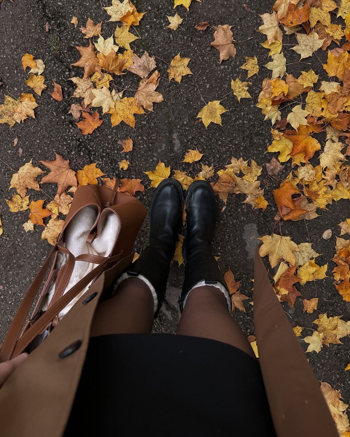 A downward view of black knee-high boots and a tan coat over autumn leaves on wet tarmac