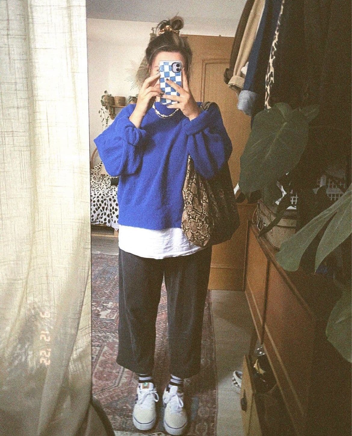 A mirror shot of someone in a cobalt blue knit jumper, white long-line tee, dark trousers and white trainers, phone raised in front of their face