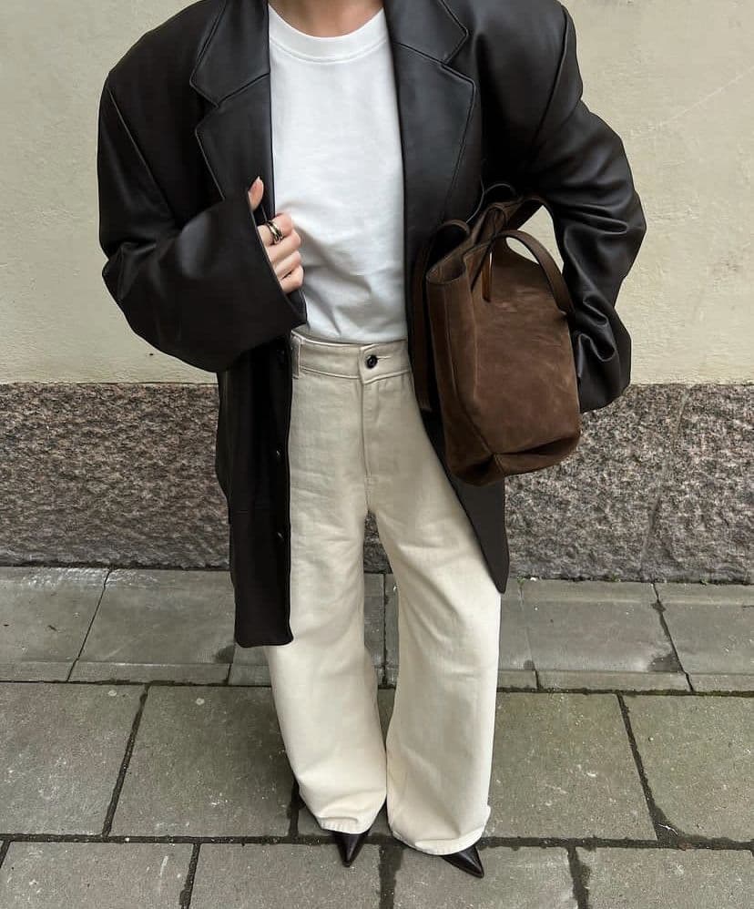 A cropped street style photo of an oversized black leather jacket over a white tee, cream wide-leg jeans, a brown suede tote and black pointed boots