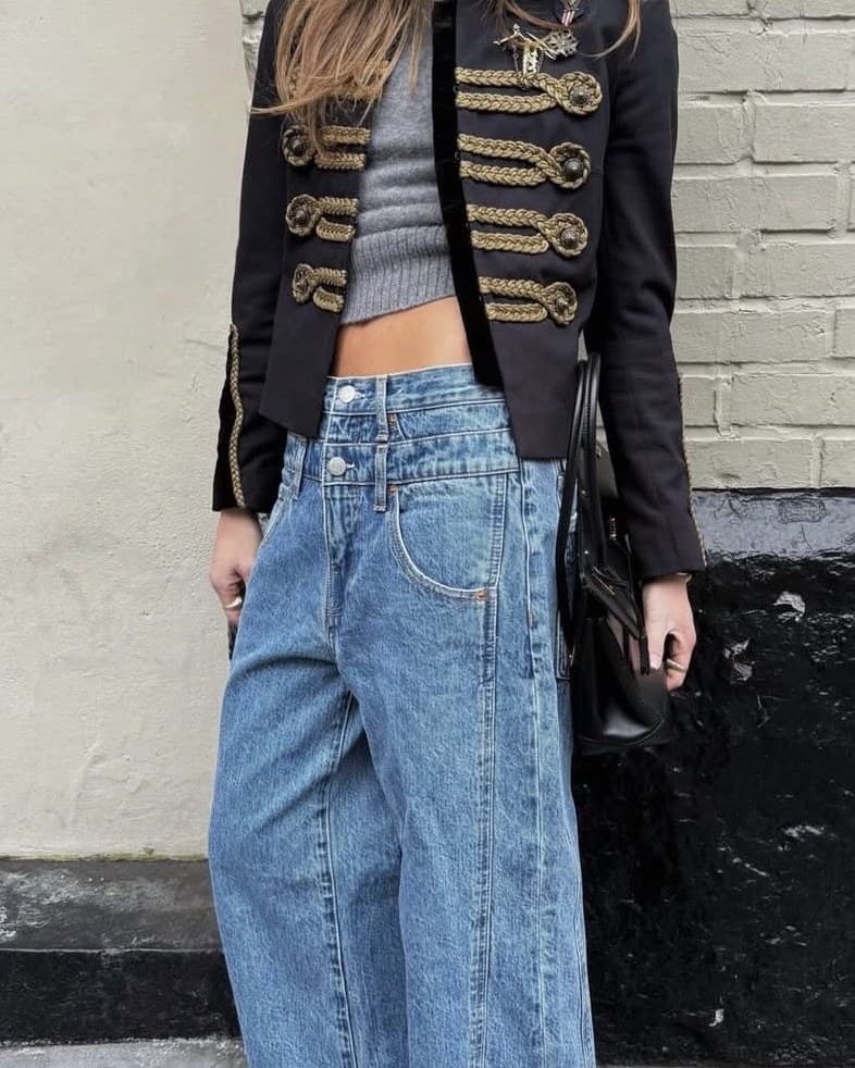 A cropped street style photo of a black military-style jacket with gold braided frogging, worn over a grey cropped ribbed knit with high-waisted wide-leg jeans