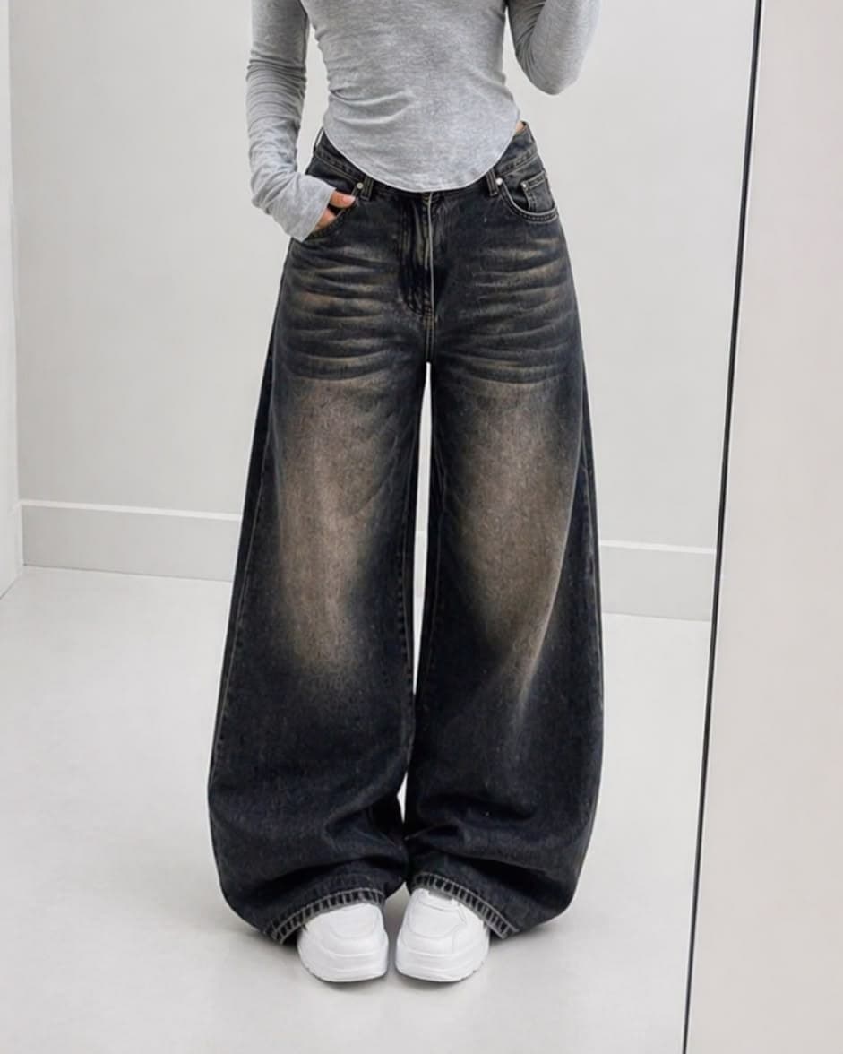 Very baggy faded black jeans with heavy sand-coloured wear on the thighs and a puddled hem, worn with a fitted grey long-sleeve top and white trainers