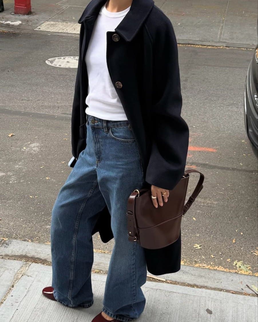 A woman photographed on a city sidewalk in a navy overcoat over a white T-shirt, wide-leg blue jeans and burgundy flats, carrying a brown leather bag