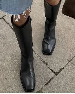 A close, cropped shot of a pair of black knee-high boots on a paving slab, isolated from the rest of the outfit