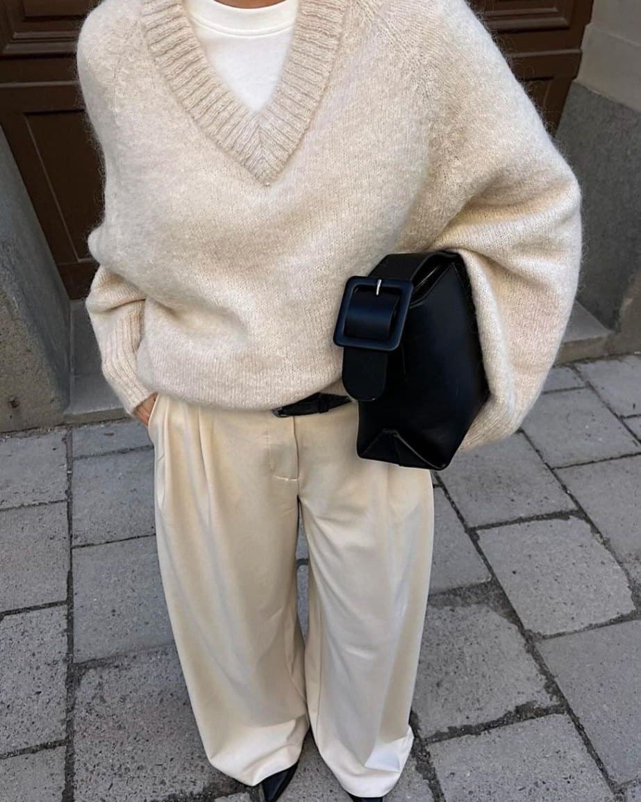 A woman photographed on a cobbled street in an oversized cream knit sweater, cream wide-leg trousers, and a black leather bag