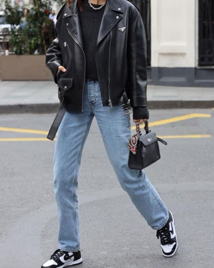 A street-style photograph cropped at the shoulders, showing an oversized black leather biker jacket over a black knit, light blue straight-leg jeans, a small black bag and black and white trainers