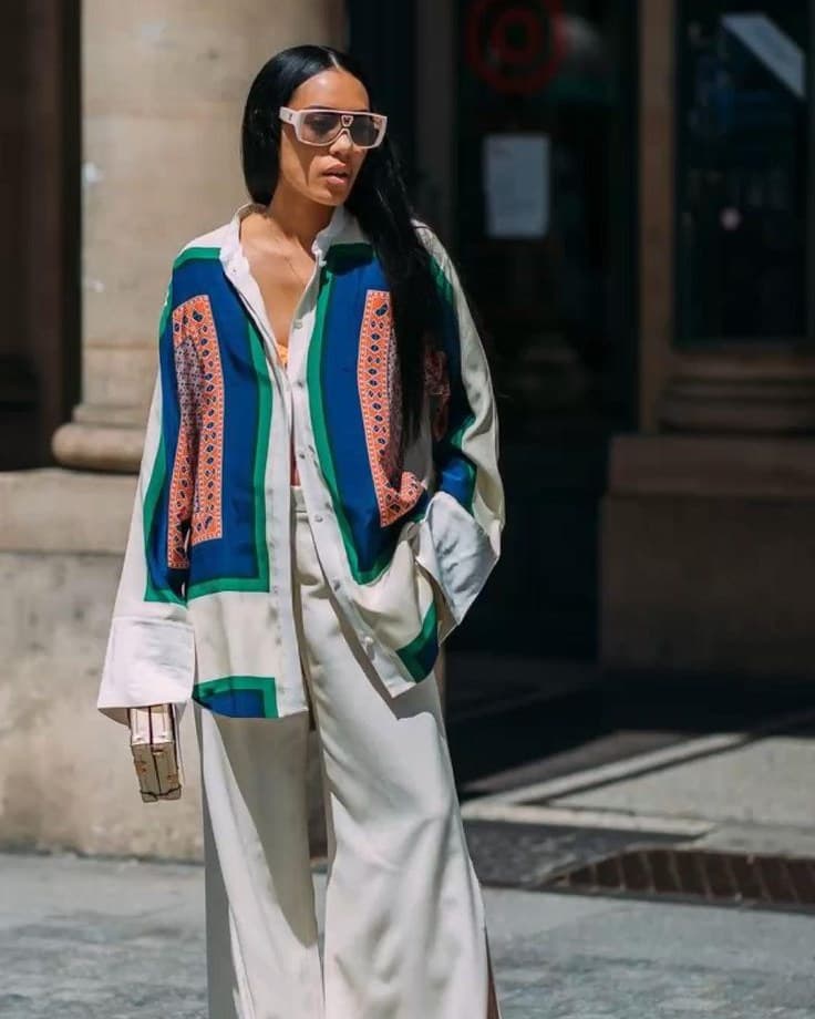 A woman photographed on a sunlit street in a patterned blue and green oversized shirt, cream wide-leg trousers and sunglasses