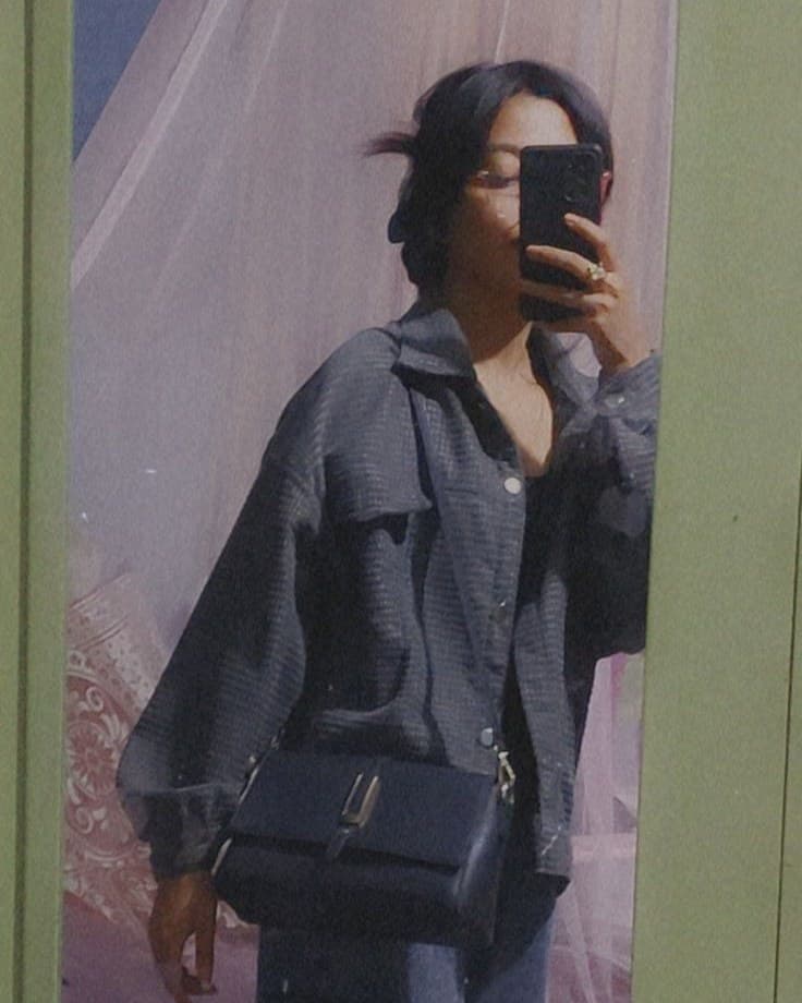 A softly compressed mirror selfie of a person in a grey-blue oversized shirt jacket with a black shoulder bag and blue jeans