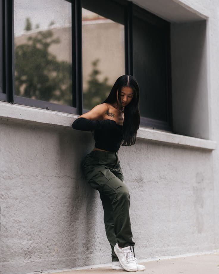 A woman leaning against a concrete wall in a black off-shoulder top, olive cargo trousers and white trainers, with mocked-up app interface chips around the edges of the frame